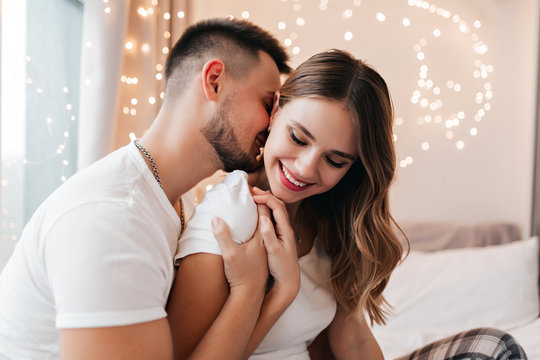 Bearded guy kissing his girlfriend in neck. Indoor romantic photoshoot of blissful young couple.