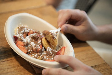 Person eating poffertjes with a plastic fork, poffertjes have chocolate, strawberries and powdered sugar on it