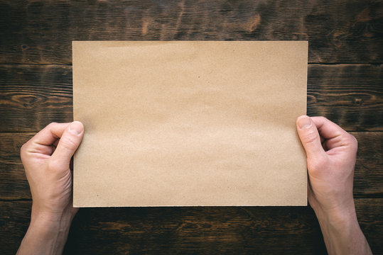 Blank brown paper page in male hands on wooden desk flat lay background.