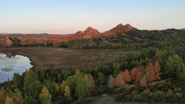 Evening autumn aerial landscape tops of rocky hills and mountains in rays of red sunset. Shore of lake or river, colored trees and shrubs, Park or reserve