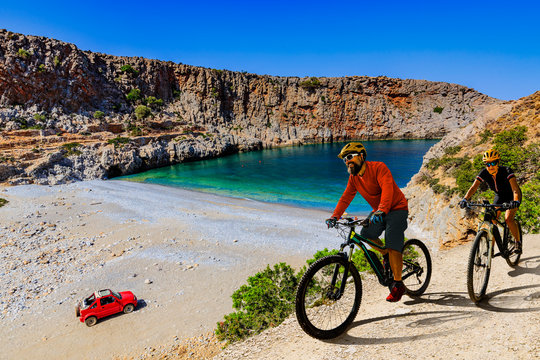 Cycling woman and man on electric bikes to wild beach with turquoise water and off-road  access . Couple cycling MTB enduro trail track. Outdoor sport activity.