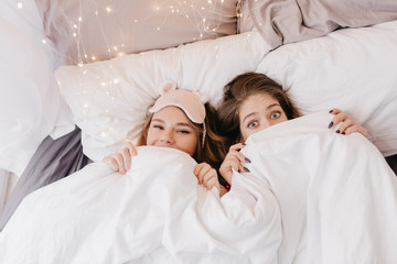 European girl with big light eyes hiding under blanket. Indoor portrait of two female models enjoying morning together.