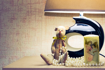 under the table lamp are a soft toy, a candle with a cartel, pearl beads