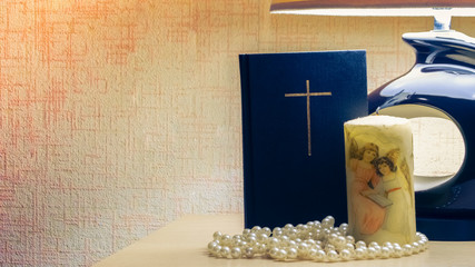 under the table lamp there is a bible, a candle with a picture, pearl beads
