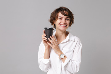Cheerful young business woman in white shirt posing isolated on grey wall background studio portrait. Achievement career wealth business concept. Mock up copy space. Hold paper cup of coffee or tea.