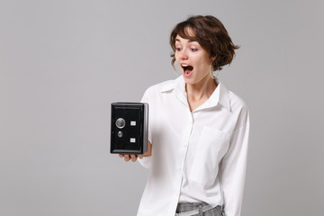 Shocked young business woman in white shirt posing isolated on grey wall background. Achievement career wealth business concept. Mock up copy space. Holding metal bank safe for money accumulation.