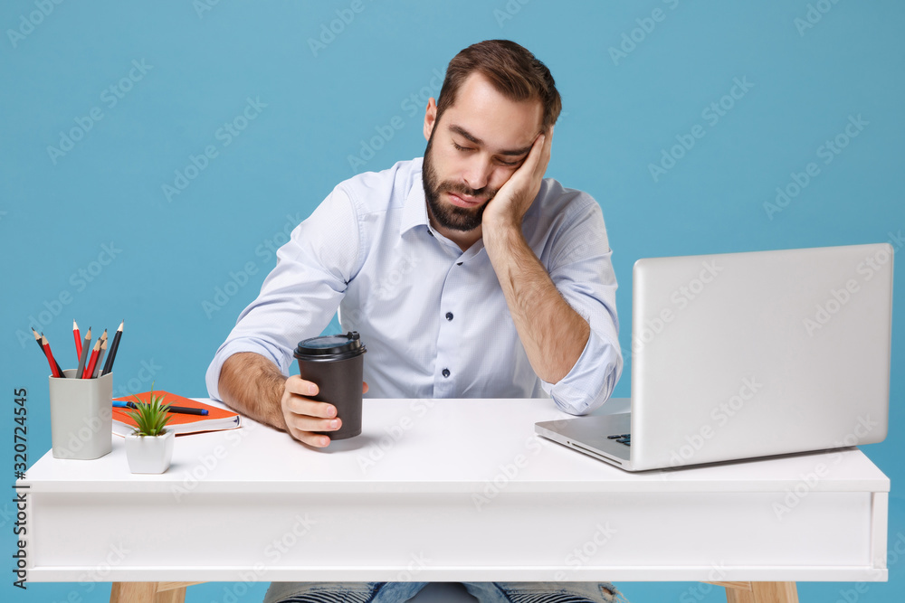 Wall mural tired young man in light shirt sit work at desk with pc laptop isolated on blue background. achievem