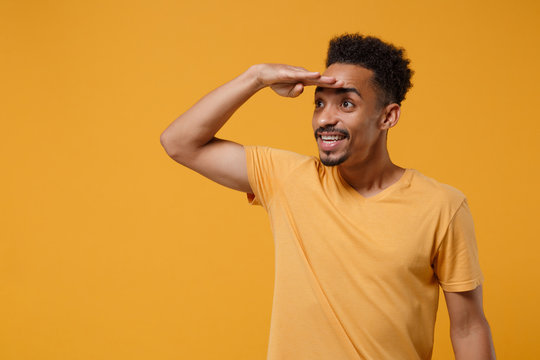 Smiling young african american guy in casual t-shirt posing isolated on yellow orange wall background. People lifestyle concept. Mock up copy space. Holding hand at forehead looking far away distance.