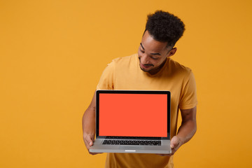 Curious young african american guy in casual t-shirt posing isolated on yellow orange wall background. People lifestyle concept. Mock up copy space. Hold laptop pc computer with blank empty screen.