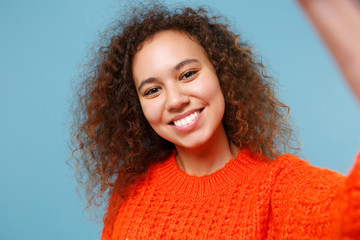 Close up of smiling young african american girl in casual orange knitted clothes isolated on pastel blue background. People lifestyle concept. Mock up copy space. Doing selfie shot on mobile phone.