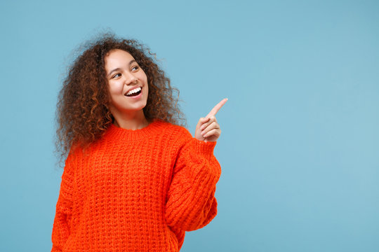 Cheerful young african american girl in casual orange knitted clothes isolated on pastel blue background studio portrait. People lifestyle concept. Mock up copy space. Pointing index finger aside up.
