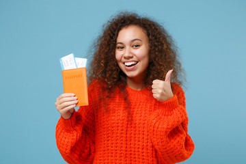 Funny young african american girl in orange knitted clothes isolated on pastel blue background. People lifestyle concept. Mock up copy space. Showing thumb up, holding passport boarding pass tickets.