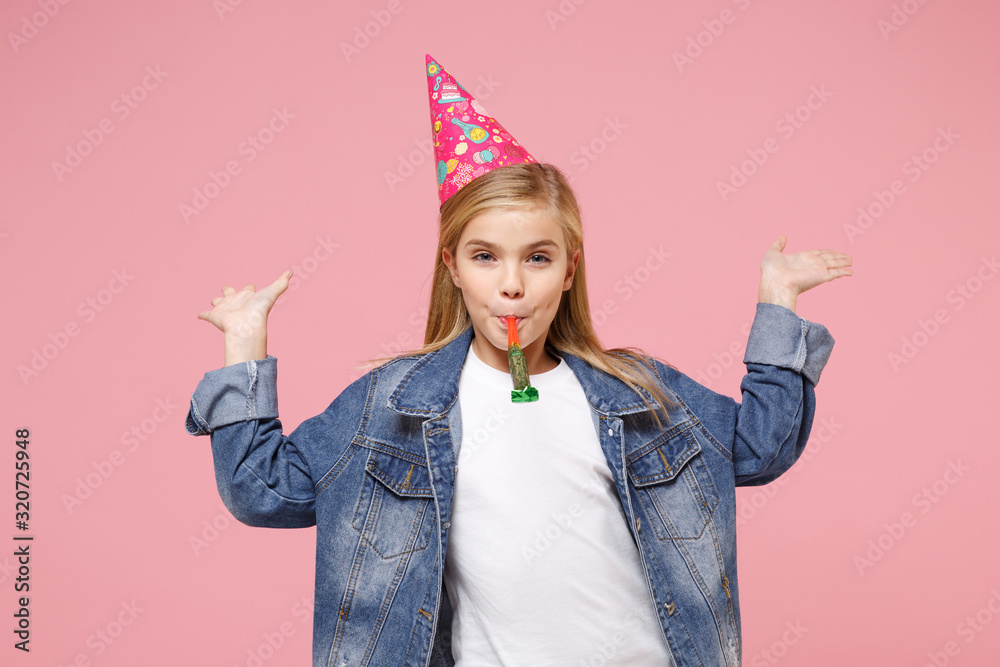 Wall mural Funny little kid girl 12-13 years old in denim jacket birthday hat isolated on pastel pink background in studio. Childhood lifestyle concept. Mock up copy space. Blowing on party pipe spreading hands.