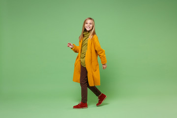 Side view of smiling little blonde kid girl 12-13 years old in yellow coat posing isolated on pastel green wall background in studio. Childhood lifestyle concept. Mock up copy space. Looking camera.