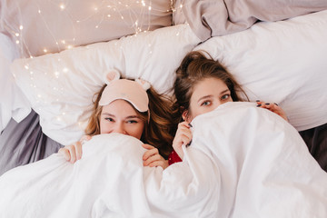 Overhead portrait of excited young ladies lying in bed. Dreamy sisters hiding under blanket early in morning.