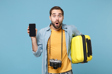 Shocked traveler tourist man in yellow clothes with photo camera isolated on blue background. Passenger traveling abroad on weekends. Air flight journey. Hold suitcase, mobile phone with empty screen.