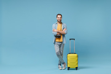 Smiling traveler tourist man in yellow clothes with photo camera, suitcase isolated on blue background. Male passenger traveling abroad on weekends. Air flight journey concept. Holding hands crossed.