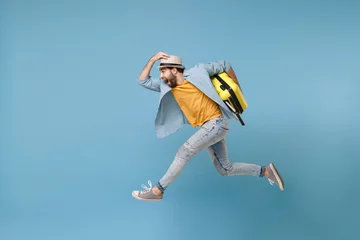 Deurstickers Side view of excited traveler tourist man in yellow clothes isolated on blue background. Male passenger traveling abroad on weekends. Air flight journey concept. Jumping like running, hold suitcase. © ViDi Studio