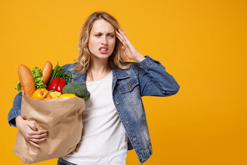 Sick young woman in denim clothes isolated on orange background. Delivery service from shop or restaurant concept. Hold brown craft paper bag for takeaway mock up with food products put hand on head.