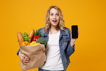 Amazed girl in denim clothes isolated on orange background. Delivery service from shop or restaurant concept. Hold brown craft paper bag for takeaway with food product mobile phone with blank screen.