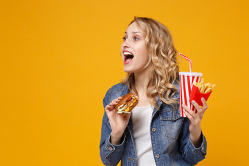 Excited young woman in denim clothes isolated on yellow orange background. Proper nutrition or American classic fast food concept. Hold burger french fries potatoes cup of cola or soda looking aside.