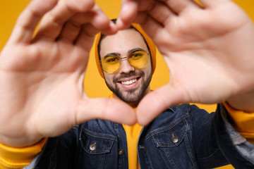 Smiling young hipster guy in fashion jeans denim clothes posing isolated on yellow orange background. People lifestyle concept. Mock up copy space. Showing shape heart with hands, heart-shape sign.