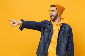 Irritated young hipster guy in fashion jeans denim clothes posing isolated on yellow orange background. People lifestyle concept. Mock up copy space. Screaming, swearing, pointing index finger aside.