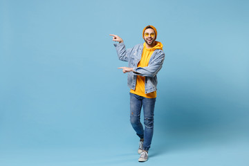 Smiling young hipster guy in fashion jeans denim clothes posing isolated on pastel blue background studio portrait. People emotions lifestyle concept. Mock up copy space. Pointing index fingers aside.