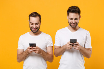 Smiling young men guys friends in white blank empty t-shirts isolated on yellow orange background studio portrait. People lifestyle concept. Mock up copy space. Using mobile phone, typing sms message.