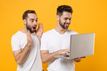 Amazed young men guys friends in white blank t-shirts posing isolated on yellow orange wall background studio portrait. People lifestyle concept. Mock up copy space. Working on laptop pc computer.