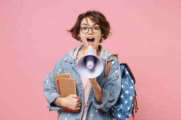 Funny young woman student in denim clothes, eyeglasses, backpack posing isolated on pastel pink background. Education in high school university college concept. Holding books, scream in megaphone.