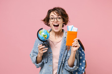 Excited young woman student in denim clothes eyeglasses, backpack posing isolated on pastel pink background. Education in high school university college concept. Hold passport tickets world globe.