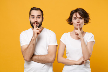 Secret couple friends bearded guy girl in white t-shirts posing isolated on yellow orange background. People lifestyle concept. Mock up copy space. Say hush be quiet with finger on lips shhh gesture.