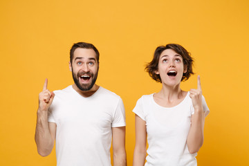 Amazed young couple friends bearded guy girl in white blank empty t-shirts posing isolated on yellow orange wall background. People lifestyle concept. Mock up copy space. Pointing index fingers up.