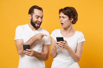 Excited couple friends bearded guy girl in white t-shirts isolated on yellow orange background. People lifestyle concept. Mock up copy space. Pointing index finger on mobile phone, typing sms message.