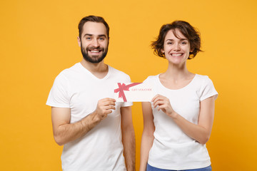 Smiling young couple friends bearded guy girl in white blank empty t-shirts isolated on yellow orange wall background in studio. People lifestyle concept. Mock up copy space. Holding gift certificate.