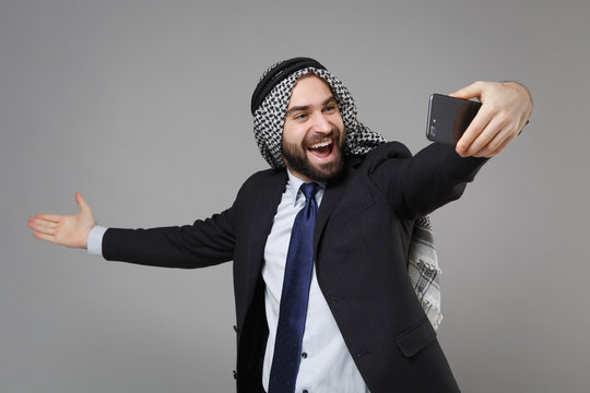 Cheerful arabian muslim businessman in keffiyeh kafiya ring igal agal suit isolated on gray background. Achievement career wealth business concept. Doing selfie shot on mobile phone spreading hands.
