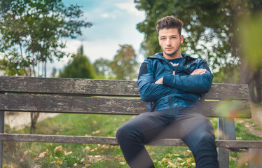 Fototapeta premium Young man in jacket sitting on bench in the park. One beautiful guy