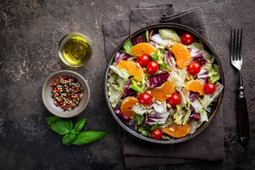 Fresh green mixed salad leaves frisee, radicchio and lamb's lettuce with tomato, olives and tangerine in a plate, top view.