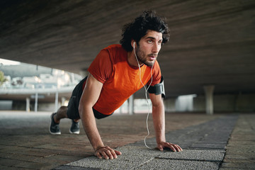 Attractive fit man listening to music in earphone doing push-ups exercises during workout outdoors...