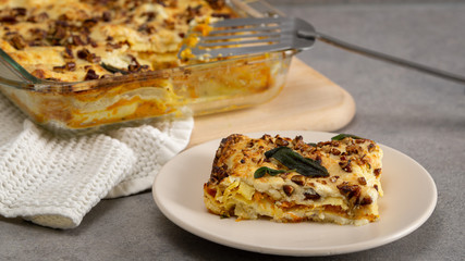 Butternut  vegetarian lasagne in a plate. With chopped nuts and herbs on it. there is whole lasagne in baking dish on a background on a wooden board. There is a cotton towel under hot dish. 