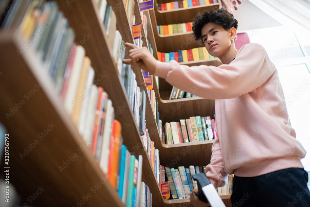 Poster teenage boy in casualwear taking book from shelf in college library while choosing one for extensive