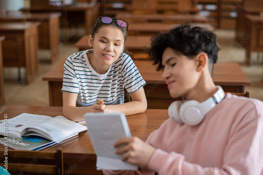 Poster Clever teenage girl pointing at notes in notepad of her classmate during discussion of points of seminar in college library
