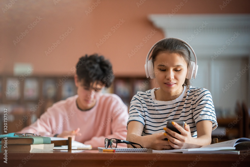 Poster Pretty college student with headphones scrolling in smartphone while sitting by desk in reading hall of college library