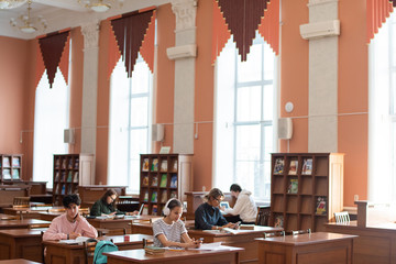Fototapeta na wymiar Two rows of desks in college library and students working individually while preparing for seminar after classes
