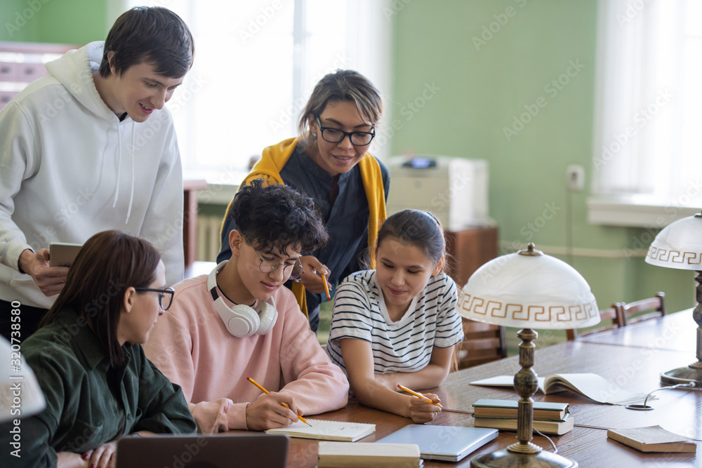 Poster Two students pointing at notes of teenage guy in his notepad while choosing name for project in group in college library