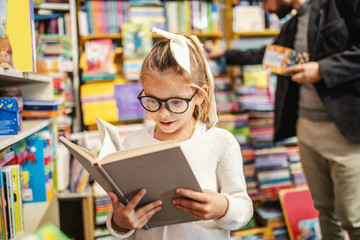 Adorable curious caucasian little girl with eyeglasses standing in bookstore and reading interesting book. All around are books on shelves.