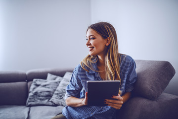 Young charming smiling caucasian brunette in stripped shirt sitting on sofa in living room, holding...