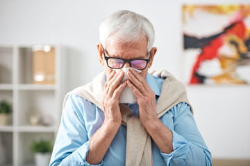 Sick senior retired man with handkerchief by his nose staying at home while feeling unwell during...