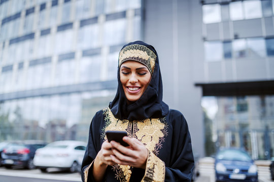 Beautiful smiling positive muslim young woman standing in front of corporate building and using smart phone for sending an e- mail. Millennial generation.